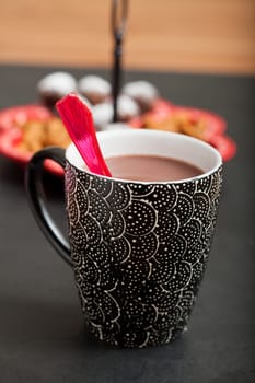 Closeup of mug of hot chocolate with red plastic spoon with tray of chocolate balls and almonds in background
