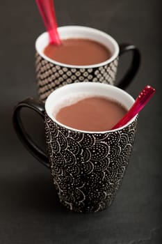 Closeup of two mugs of hot chocolate with red plastic spoons on dark surface