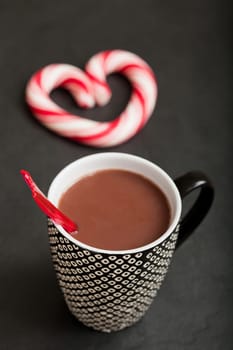 Closeup of mug of hot chocolate with red plastic spoon and hard candy heart with red swirls in background