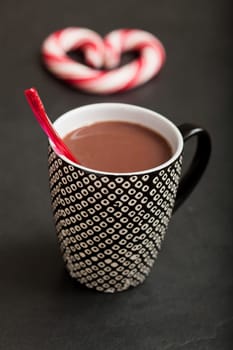 Closeup of mug of hot chocolate with red plastic spoon and hard candy heart with red swirls in background