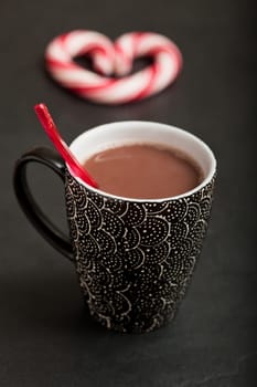 Closeup of mug of hot chocolate with red plastic spoon and hard candy heart with red swirls in background