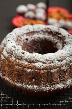 Closeup of dry chocolate cake sprinkled with icing sugar resting on metal grill with tray of chocolate balls and almonds in background
