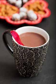 Closeup of mug of hot chocolate with red plastic spoon with tray of chocolate balls and almonds in background