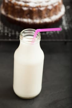 Closeup of milk in glass jar with pink straw and chocolate cake sprinkled with icing sugar resting on metal grill in background
