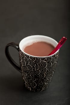 Closeup of mug of hot chocolate with red plastic spoon on dark surface