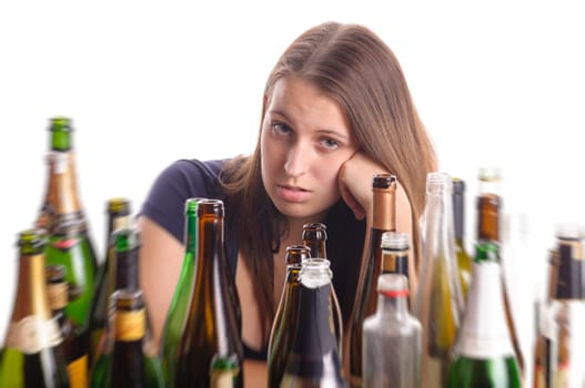 A young woman with brunette long hair sits at a table in the middle of empty bottles and looks desperate,isolated against a white background.