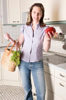 Adult housewife stands with a basket full of fresh vegetables in her bright fitted kitchen.