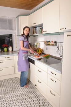 Adult housewife stands in her bright fitted kitchen and prepares fresh vegetables for cooking.