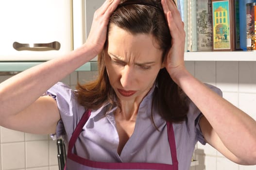 Adult housewife stands in her bright fitted kitchen and holds her hands to her head.