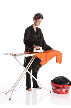 Adult housewife with curlers in her hair and a bathrobe stands behind the ironing board and irons with the iron against a white background.