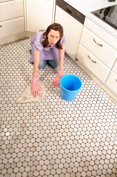 Adult housewife kneels in her bright fitted kitchen and cleans the floor sour and frustrated with bucket and cleaning cloth.