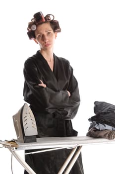 Adult housewife with curlers in her hair and a bathrobe stands behind the ironing board and irons with the iron against a white background.
