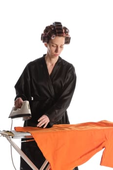 Adult housewife with curlers in her hair and a bathrobe stands behind the ironing board and irons with the iron against a white background.
