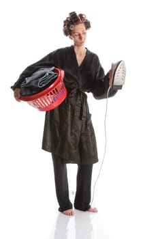 Adult housewife with curlers in her hair and a bathrobe stands against a white background and with a basket of ironing and an iron.