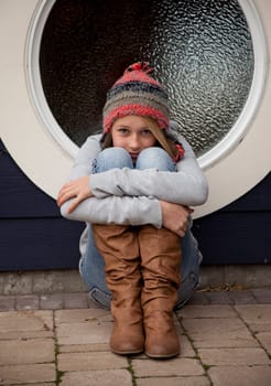Outdoor photo of young teenage girl seated on ground, hugging knees, distraught facial expression.