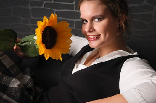 A young girl with a blouse and boots sits teasingly on a black leather armchair against a dark background and plays with a sunflower