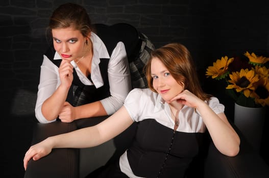 Two young women (friends) sit together in a black leather armchair against a dark background