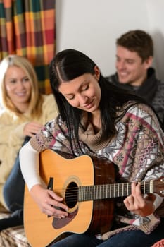 Young friends in winter cottage play guitar spend holiday break