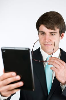 Upper body shot of a young man in a black suit and glasses against a white background looking towards a tablet computer held in front of him in the direction of the camera