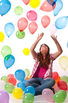 Teenage girl sits on the floor and stretches her hands up to catch colorful balloons against a white background