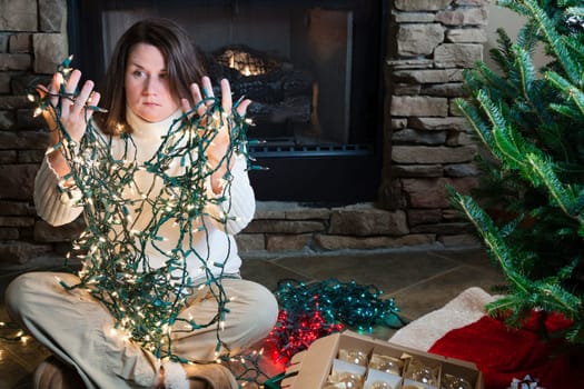 Young woman sitting next to Christmas tree holding entangled string of lights