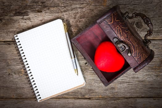 Red heart in the  box and notebook with pen on wooden floor