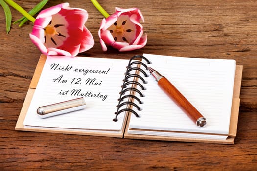 Notebook opened with the text "Don't forget! May 12 is Mother's Day"" and a pen lies on wooden ground"