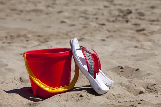 Flip flops leaning against a red bucket stand on the beach