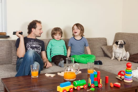 father with sons and dog on sofa, in the foreground on a table toys and drinks