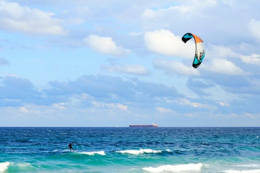 FORT LAUDERDALE, FLORIDA - FEBRUARY 9, 2013: A kiteboarder sailing off the blue green Atlantic ocean coast in the morning with a red orange tanker cargo ship traveling far off in the background.