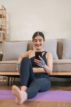 Lifestyle asian young fitness woman holding smartphone relaxing after workout at home. Smiling female using cell phone checking newsfeed on social media while exercise.