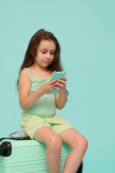 Adorable little child girl using smartphone while sitting on her suitcase, awaiting her flight, checking mobile app, isolated over blue color background. Copy ad space. Kids. Travel. Technology
