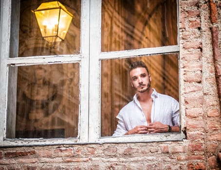 A man standing in front of a window. Photo of a confident man with hands in pockets looking out of a window