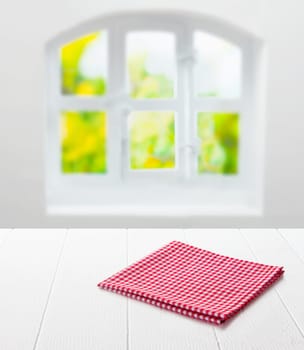 Red and white checked cloth neatly folded on top of an empty clean white table top under a window with a view of summer greenery in a country kitchen