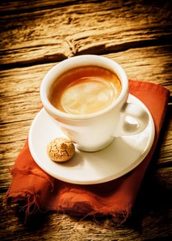 Cup of hot frothy espresso coffee served on a rustic cloth with a small macaroon in a rural country cafeteria on a weathered wood surface with vignetting and copyspace