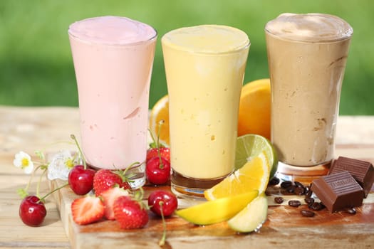 Three delicious smoothies with yoghurt or ice cream blend, two made with fruit and one of chocolate, together with various fresh tropical fruit on a garden table