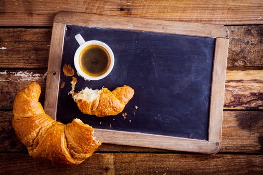 Overhead view of a fresh cup of coffee and a flaky croissant broken in two on an old school slate over a rustic wooden background, copyspace on the slate