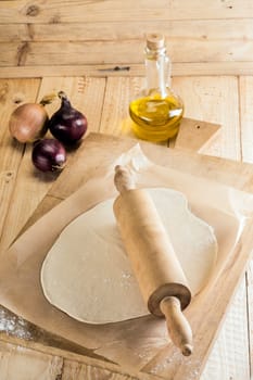 Ingredients for Italian pizza with freshly rolled dough for the pie crust or base on a wooden board with a rolling pin, onions and a decanter of olive oil
