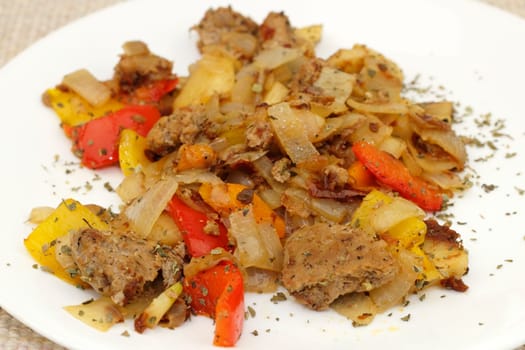 Orange carrots, white onions, turkey, red, orange, yellow sweet peppers, sun dried tomatoes and oregano on a round white plate in front of a cloth brown place mat background.