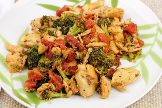 Chunks of boneless chicken cooked mixed with cut tomatoes, broccoli and oregano and set on a round white plate that has bamboo leaves painted on it placed on a brown cloth place mat..