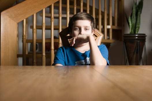 Frontal torso view of a young male teenager in a blue T-shirt sitting bored at a wooden table and looking at the camera with his hand held to his mouth