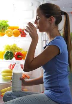 Young woman drinking milk near refrigerator at night