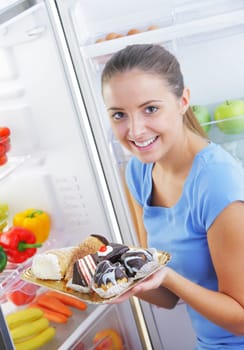 Beautiful young woman sitting near refrigerator  and choosing between healthy fruits and tasty cakes