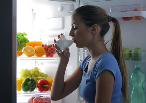 Young woman drinking milk near refrigerator at night