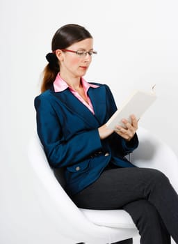 Young woman in a suit and glasses reading a book while sitting in a chair