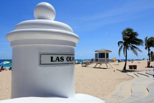 FORT LAUDERDALE, FLORIDA - APRIL 8, 2013: Entrance sign to Las Olas beach looking south along the Atlantic ocean coast with lifeguard tower 6 and many people relaxing along its sunny tropical shore.