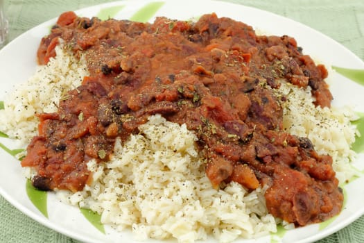 Closeup of vegetarian dinner of organic oregano herb, diced tomatoes, kidney beans, black beans and pinto over brown rice on a round white plate with painted bamboo leaves placed on a green placemat.