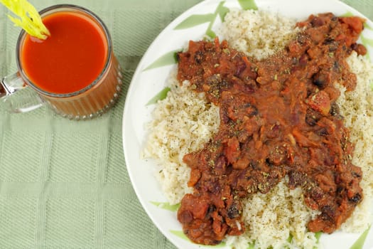 White dinner plate of brown rice topped with organic black, pinto and red kidney beans, diced tomato, oregano, and pepper. Drink is a clear mug of vegetable juice with a celery heart stalk garnish..