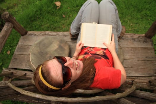 A freckled woman wearing sunglasses smiles up and back at the camera while reading a book on an unique wooden bench.
