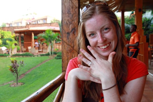 A smiling woman sits in a tropical place displaying her simple and cheap engagement ring made out of string.